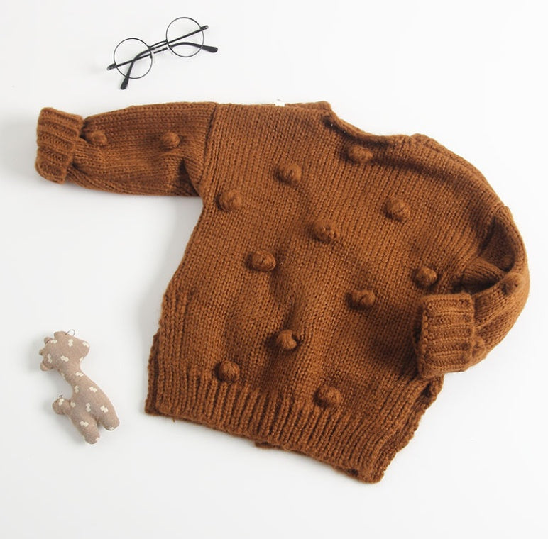 Brown knitted cute toddler cardigan for girls with pompom details laid flat next to glasses and toy