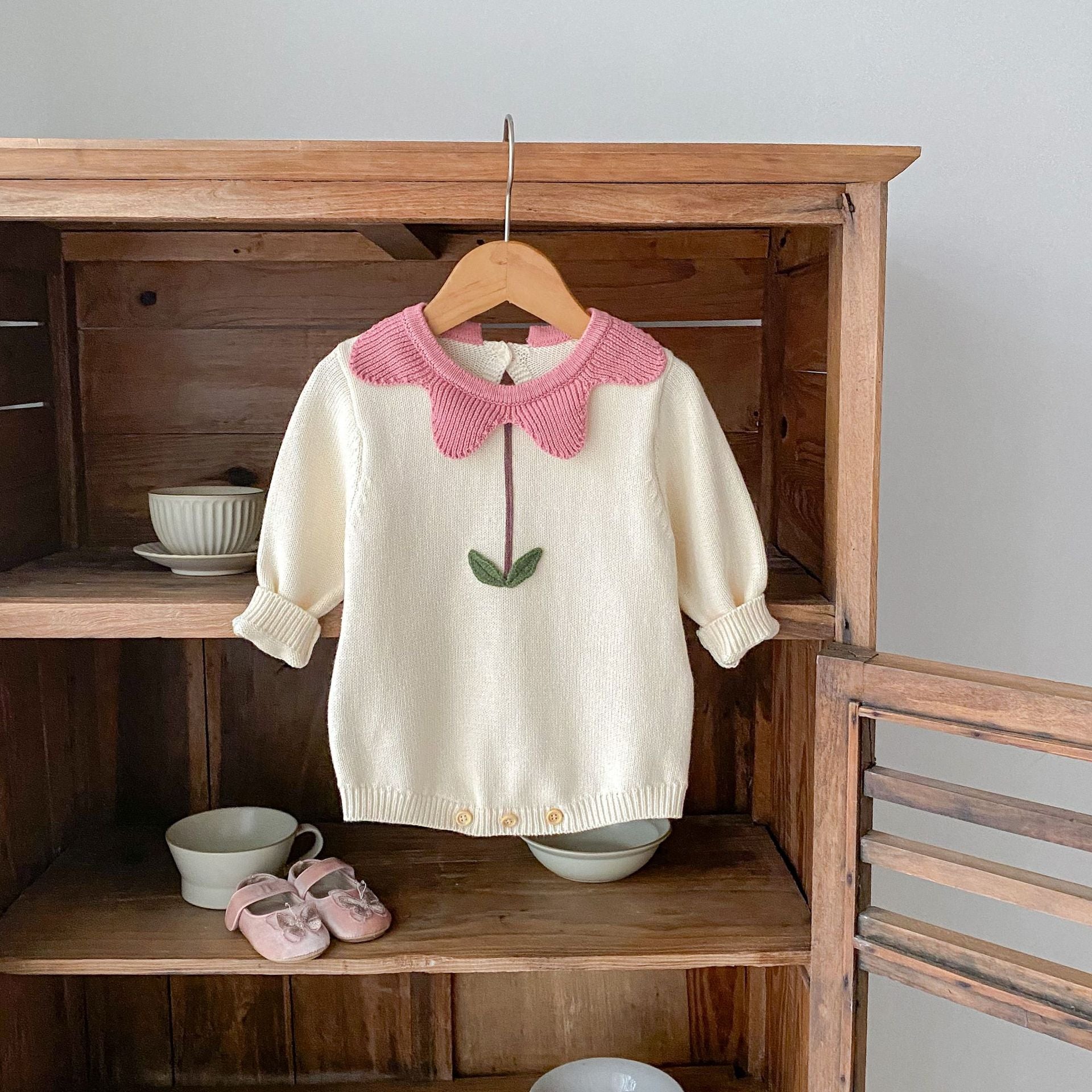White floral newborn sweater with pink petal collar and green leaf detail hanging on wooden hanger