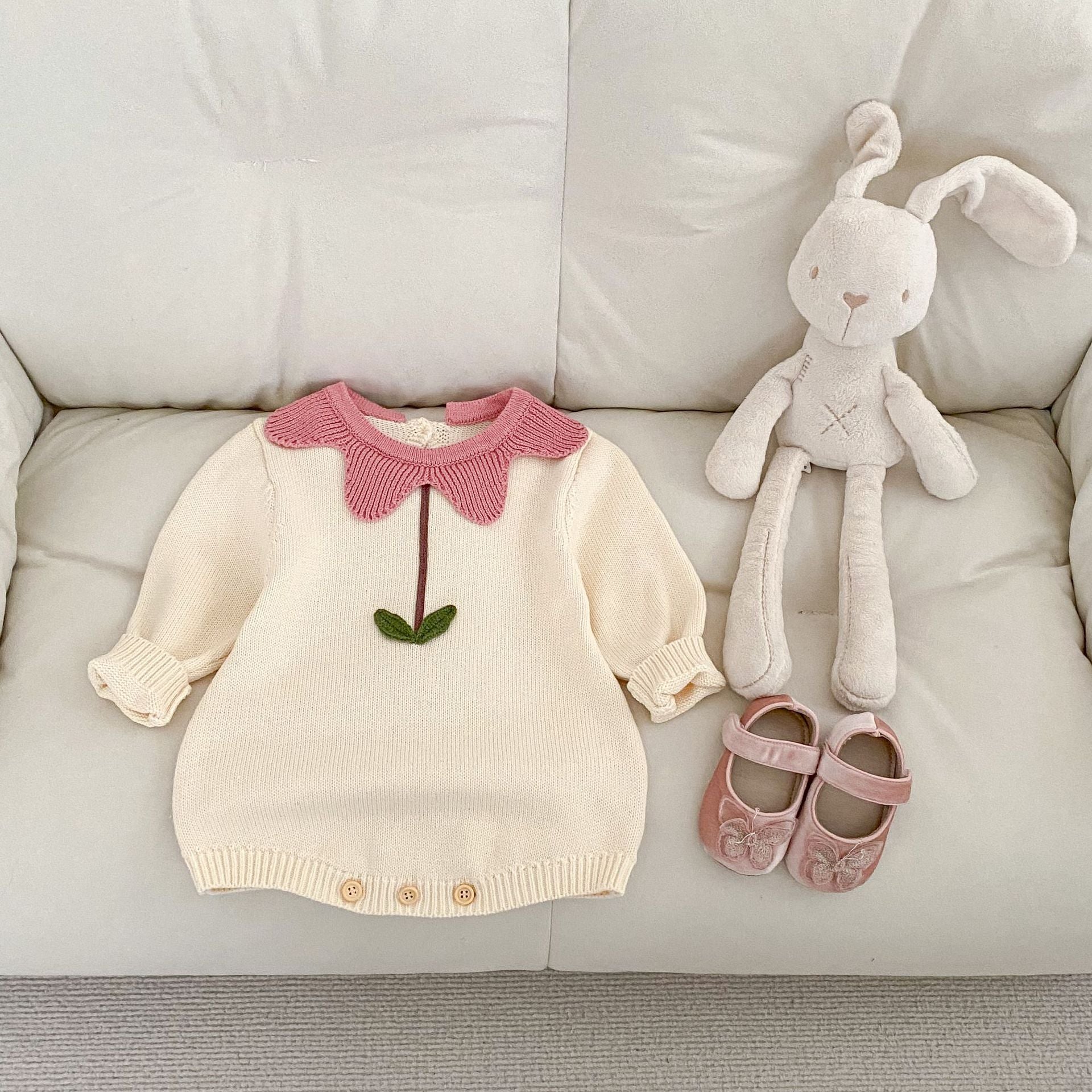 Cream-colored floral newborn sweater with pink collar and green leaf detail laid on couch with plush bunny and pink baby shoes
