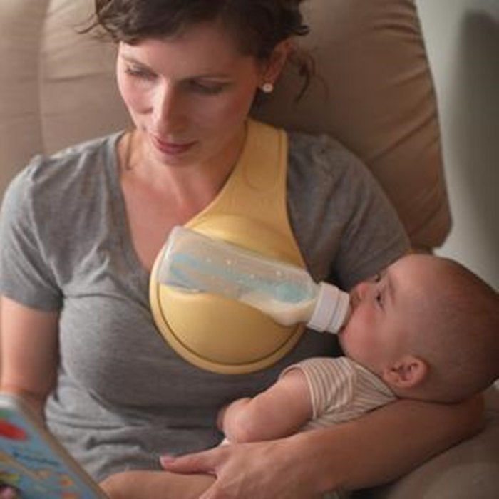 Mother feeding baby with hands free baby bottle holder while reading a book in a cozy chair