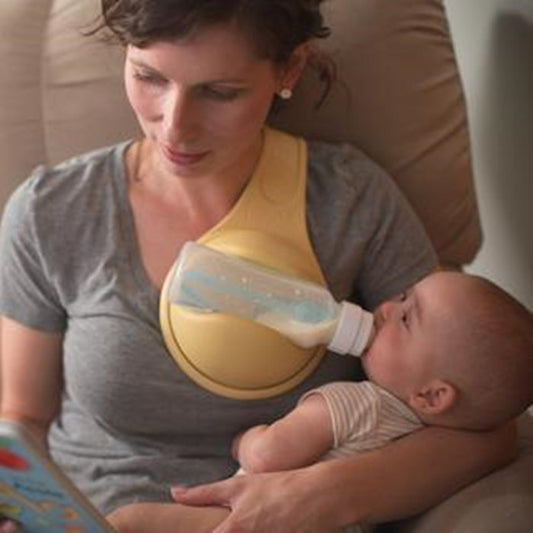 Mother feeding baby with hands free baby bottle holder while reading a book in a cozy chair