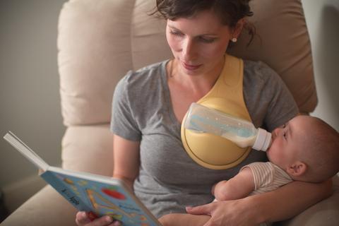 Mother using hands free baby bottle holder to feed infant while reading a book in a chair