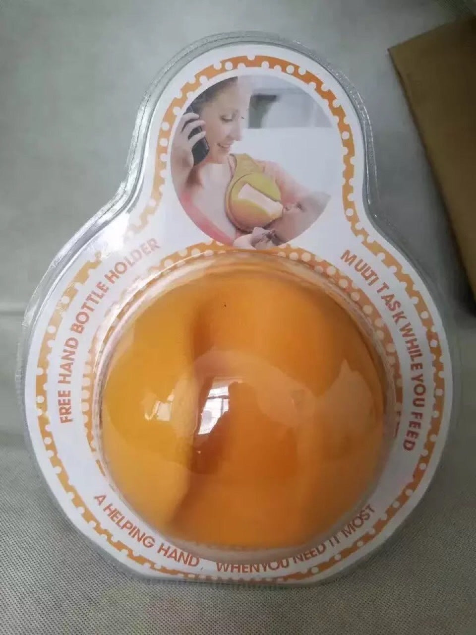 Hands free baby bottle holder in orange with packaging showing a mother multitasking while feeding baby